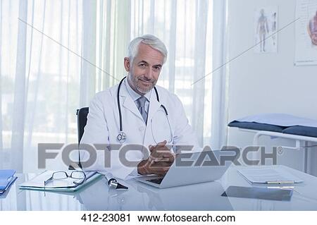 Portrait of smiling mature doctor sitting at desk with laptop in office View Large Photo Image Stock Image - Portrait of smiling mature doctor sitting at desk with laptop in office. Fotosearch