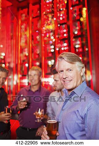 Stock Photography - Portrait of smiling mature man holding beer glass in nightclub, friends in background. Fotosearch