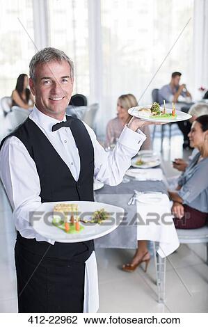 Portrait of smiling waiter holding fancy dishes, people sitting at restaurant tables in background View Large Photo Image Stock Image - Portrait of smiling waiter holding fancy dishes, people sitting at restaurant tables in background. Fotosearch
