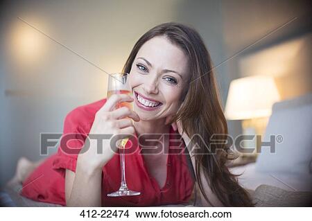 Picture - Portrait of smiling woman lying on bed with champagne flute. Fotosearch