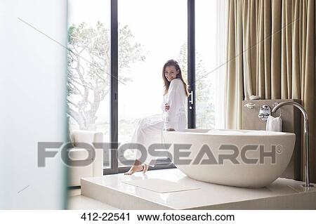 Portrait of smiling woman wearing white bathrobe, sitting on edge of bathtub in bathroom View Large Photo Image Stock Image - Portrait of smiling woman wearing white bathrobe, sitting on edge of bathtub in bathroom. Fotosearch