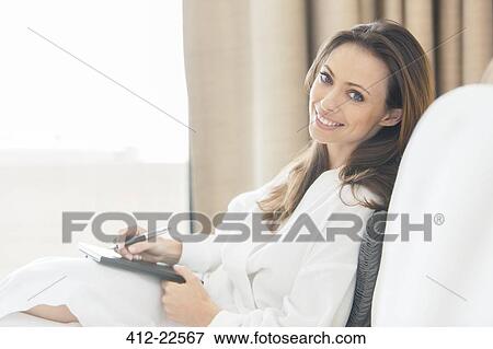 Stock Photo - Portrait of smiling woman wearing white bathrobe with notebook in bedroom. Fotosearch