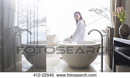 Stock Photography - Portrait of smiling woman wearing white bathrobe, sitting on edge of bathtub in bathroom. Fotosearch