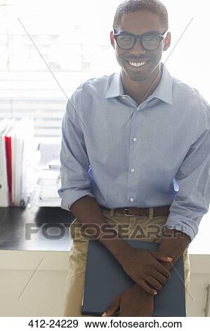 Portrait of smiling young man wearing glasses and blue shirt leaning on desk in office View Large Photo Image Stock Photo - Portrait of smiling young man wearing glasses and blue shirt leaning on desk in office. Fotosearch