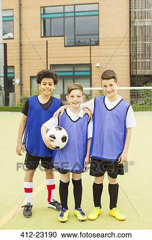 Stock Image - Portrait of three smiling boys wearing soccer uniforms and holding soccer ball in front of school. Fotosearch