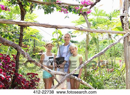 Stock Photo - Portrait of woman and two girls standing in garden with shovel and tree seedling. Fotosearch