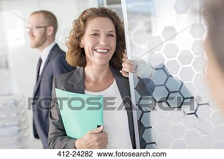 Portrait of woman holding documents standing at glass door, colleague in background View Large Photo Image Stock Image - Portrait of woman holding documents standing at glass door, colleague in background. Fotosearch
