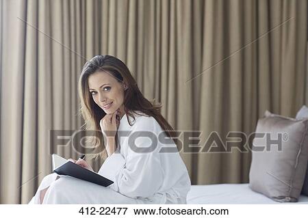 Stock Photo - Portrait of woman wearing bathrobe with notepad in bedroom. Fotosearch