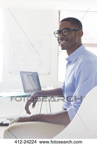 Picture - Portrait of young smiling man wearing glasses and blue shirt sitting at desk with laptop. Fotosearch