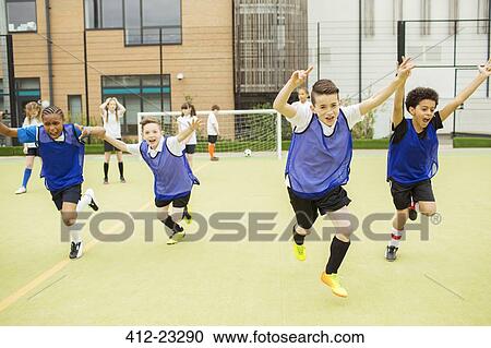 Stock Image - Schoolboys wearing sport uniforms running with arms raised in soccer field in front of school. Fotosearch