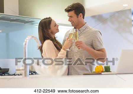Smiling couple holding champagne flutes and looking at each other in their kitchen View Large Photo Image Stock Photo - Smiling couple holding champagne flutes and looking at each other in their kitchen. Fotosearch