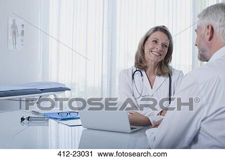 Stock Image - Smiling female doctor talking to patient at desk in office. Fotosearch