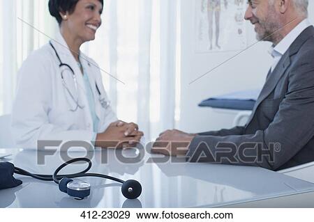 Smiling female doctor talking to patient at desk in office, blood pressure gauge in foreground View Large Photo Image Stock Photo - Smiling female doctor talking to patient at desk in office, blood pressure gauge in foreground. Fotosearch