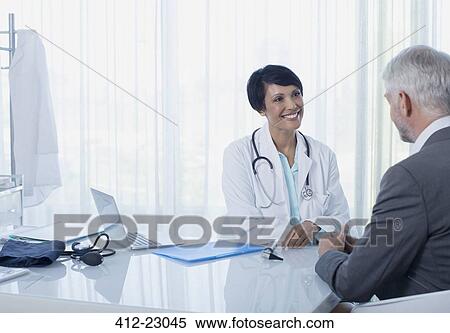 Stock Photography - Smiling female doctor talking to patient at desk in office. Fotosearch
