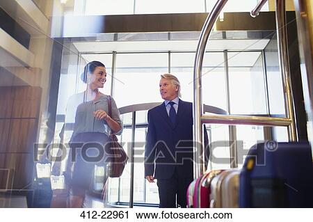 Stock Image - Smiling man and woman entering hotel lobby, suitcases on luggage cart in foreground. Fotosearch