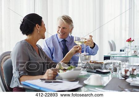 Stock Image - Smiling mature couple raising toast with white wine at restaurant table. Fotosearch