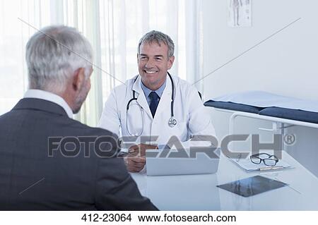 Smiling mature doctor and man sitting at desk in office View Large Photo Image Picture - Smiling mature doctor and man sitting at desk in office. Fotosearch