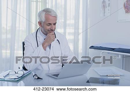 Smiling mature doctor sitting at desk with laptop in office View Large Photo Image Picture - Smiling mature doctor sitting at desk with laptop in office. Fotosearch