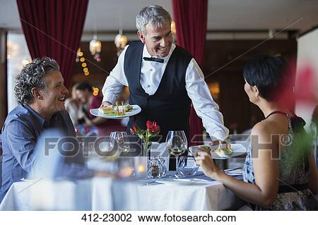 Smiling waiter serving fancy dishes to mature couple sitting at table in restaurant View Large Photo Image Stock Image - Smiling waiter serving fancy dishes to mature couple sitting at table in restaurant. Fotosearch