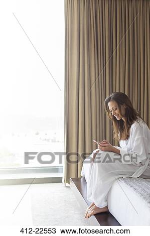 Stock Image - Smiling woman in bathrobe texting in bedroom. Fotosearch