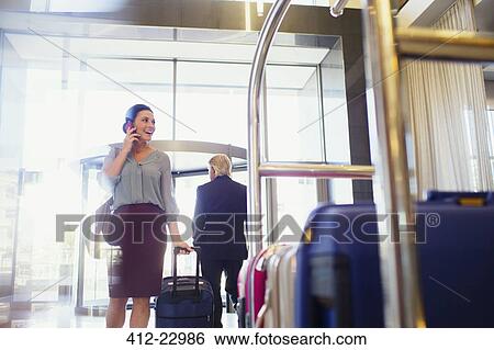 Stock Photograph - Smiling woman talking on phone in hotel lobby, luggage cart in foreground. Fotosearch