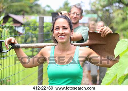 Stock Photo - Smiling woman wearing green tank top carrying shovel on shoulders through community garden. Fotosearch