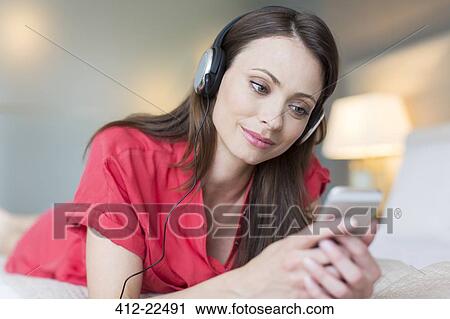 Stock Image - Smiling woman wearing red dress lying on bed listening to music from smartphone. Fotosearch
