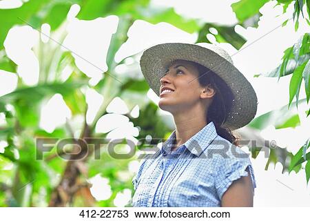 Smiling woman wearing straw hat looking at plants in sunny garden View Large Photo Image Stock Image - Smiling woman wearing straw hat looking at plants in sunny garden. Fotosearch