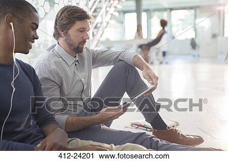 Two businessmen using digital tablet and earphones sitting on floor in modern office View Large Photo Image Stock Image - Two businessmen using digital tablet and earphones sitting on floor in modern office. Fotosearch