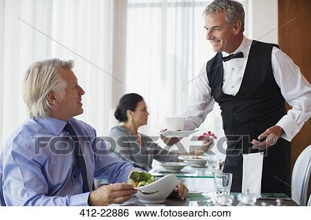 Stock Photograph - Waiter serving cup of coffee to mature man sitting at table in restaurant. Fotosearch