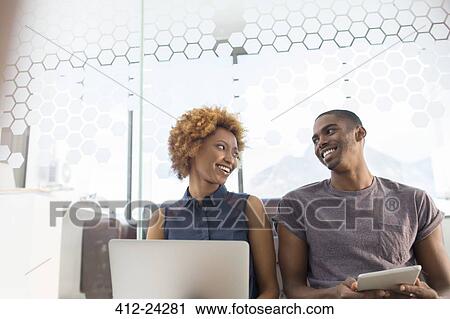 Stock Image - Woman using laptop and man using tablet smiling, looking at each other. Fotosearch