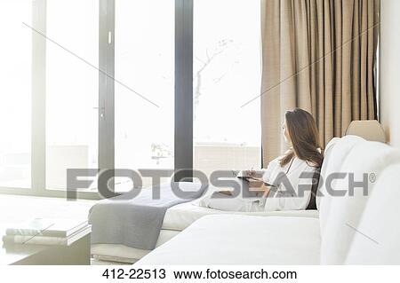Stock Image - Woman wearing white bathrobe reclining on sofa in living room and looking through window. Fotosearch