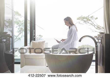 Woman wearing white bathrobe sitting on edge of modern bathtub and using digital tablet View Large Photo Image Stock Photo - Woman wearing white bathrobe sitting on edge of modern bathtub and using digital tablet. Fotosearch