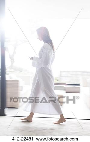 Stock Image - Woman wearing white bathrobe walking through modern corridor with mobile phone. Fotosearch