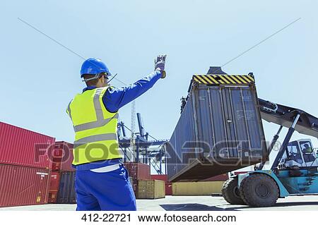 Stock Image - Worker directing crane carrying cargo container. Fotosearch