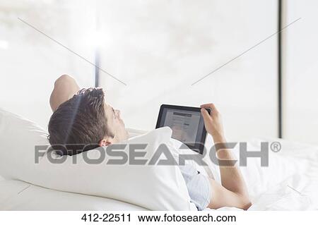 Young man lying on bed and using tablet View Large Photo Image Stock Image - Young man lying on bed and using tablet. Fotosearch