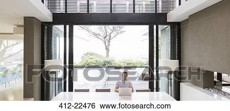 Young man using laptop in modern dining room with patio doors and swimming pool in background View Large Photo Image Stock Photograph - Young man using laptop in modern dining room with patio doors and swimming pool in background. Fotosearch