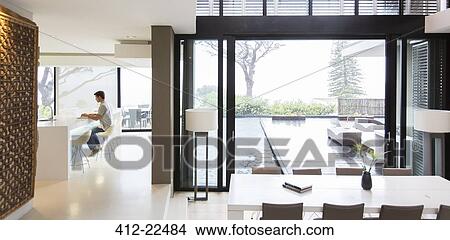 Picture - Young man using laptop in modern kitchen, dining area with patio door and swimming pool in foreground. Fotosearch