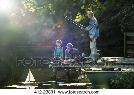 Stock Image - Boy fishing and playing with toy sailboat with father and grandfather at lake. Fotosearch