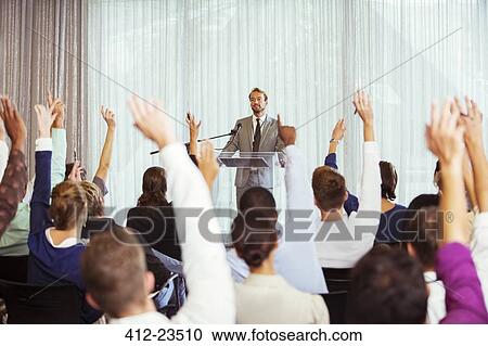 Stock Image - Businessman giving presentation in conference room, people raising hands. Fotosearch