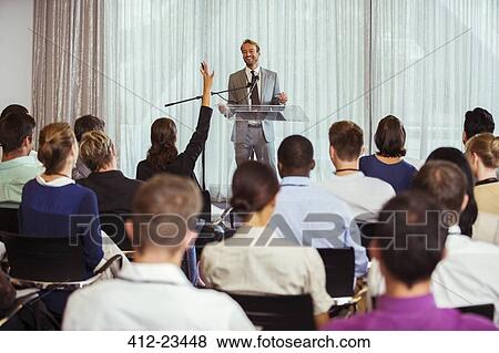 Stock Photo - Businessman giving speech in conference room, woman from audience raising hand. Fotosearch