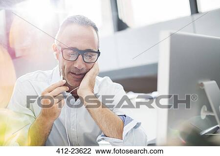 Stock Image - Businessman listening to earbuds at office desk. Fotosearch