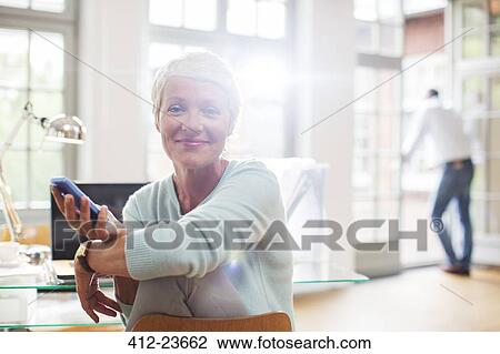 Stock Image - Businesswoman using cell phone at home office desk. Fotosearch