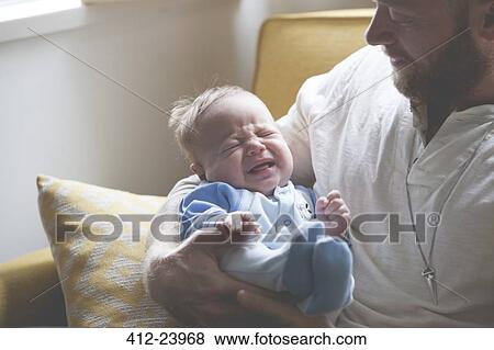 Stock Photo - Father holding and looking at crying baby, sitting on sofa. Fotosearch