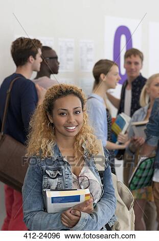 Stock Photograph - Female student holding books and smiling at camera with group of students in background. Fotosearch