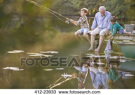 Stock Image - Grandfather and grandsons fishing and playing with toy sailboat at lake. Fotosearch