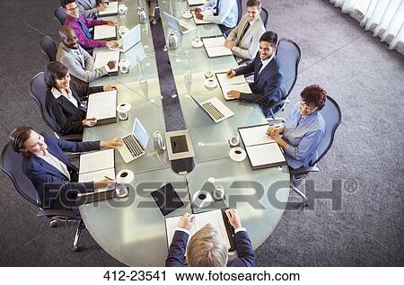 Stock Image - Group of businesspeople sitting at conference table looking at chairman. Fotosearch