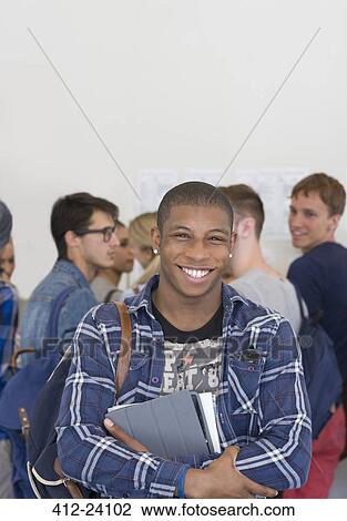Stock Image - Male student holding books and smiling at camera with other students in background. Fotosearch
