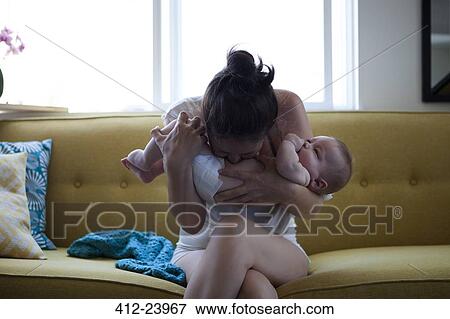 Stock Photo - Mother holding and kissing baby's belly, sitting on sofa by window. Fotosearch