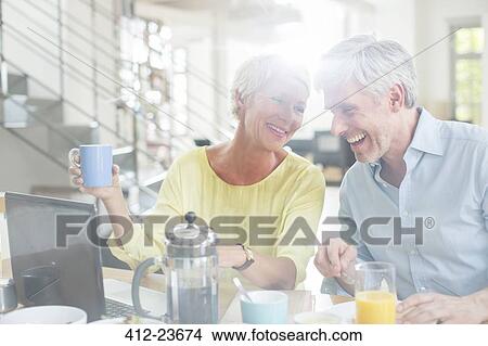Picture - Older couple laughing together at breakfast table with laptop. Fotosearch